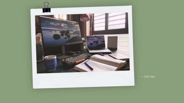 What's on my desk at seven in the morning: monitor, laptop, keyboard, mug, an open book and a notebook in early light