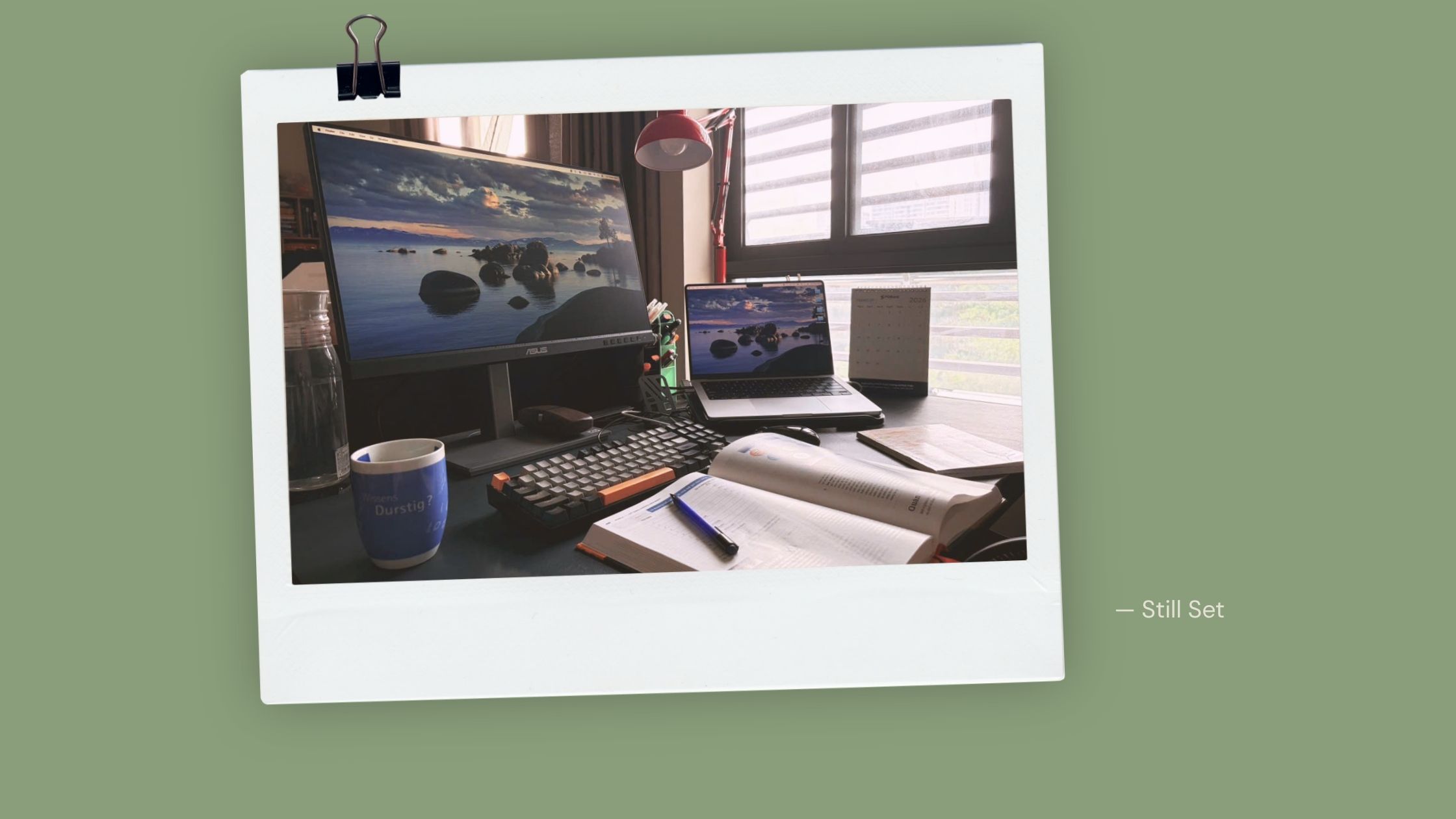 What's on my desk at seven in the morning: monitor, laptop, keyboard, mug, an open book and a notebook in early light